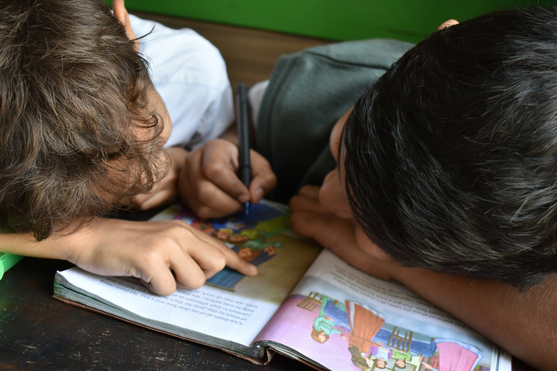 Two kids lay their head on a book and talk to eachother.