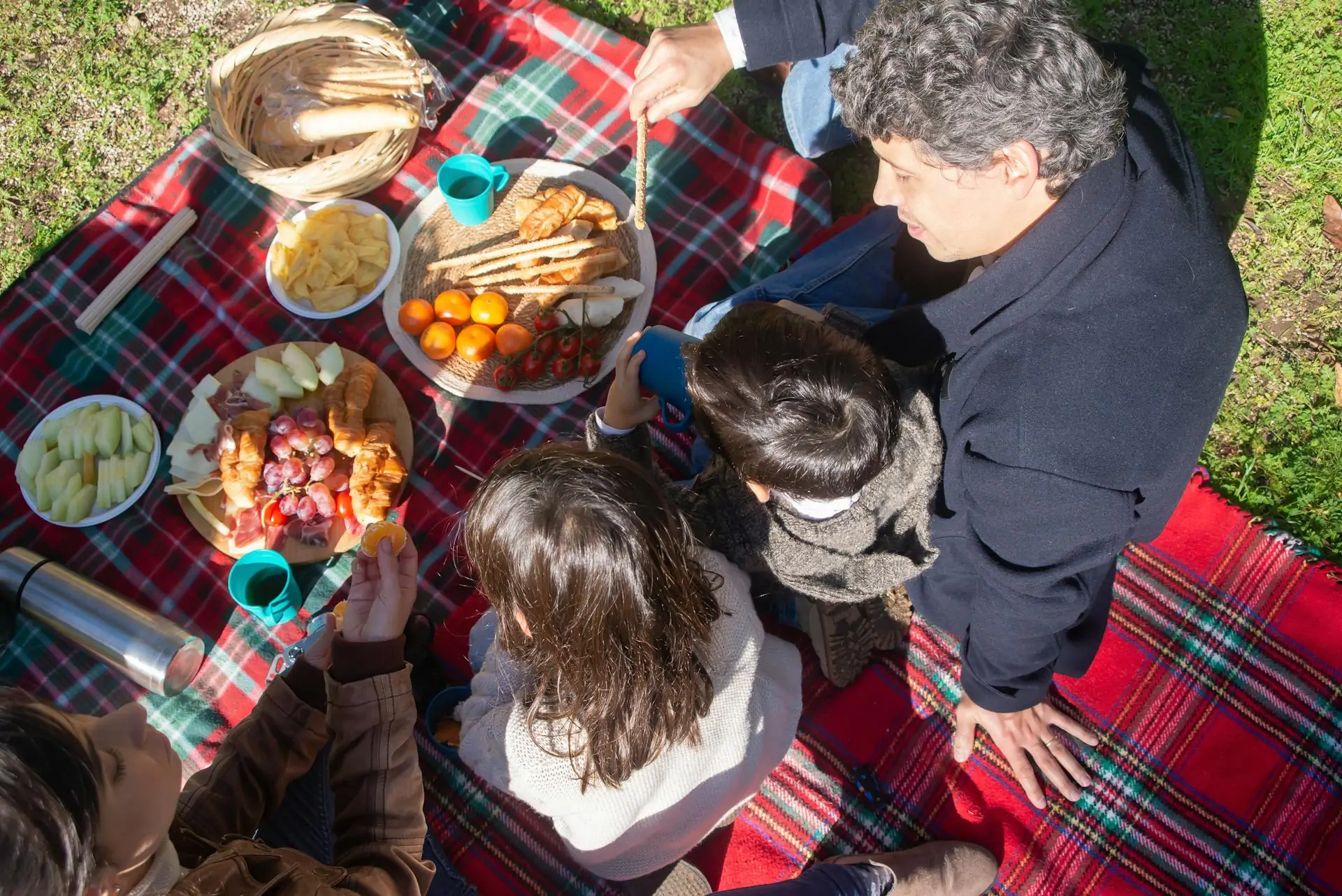 Parent sits with two children on a picnic blanket with a variety of food set up on the blanket.