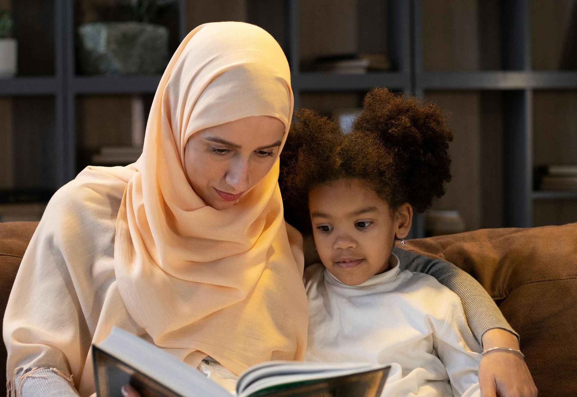 A mother sits by her daughters side and reads her a book.